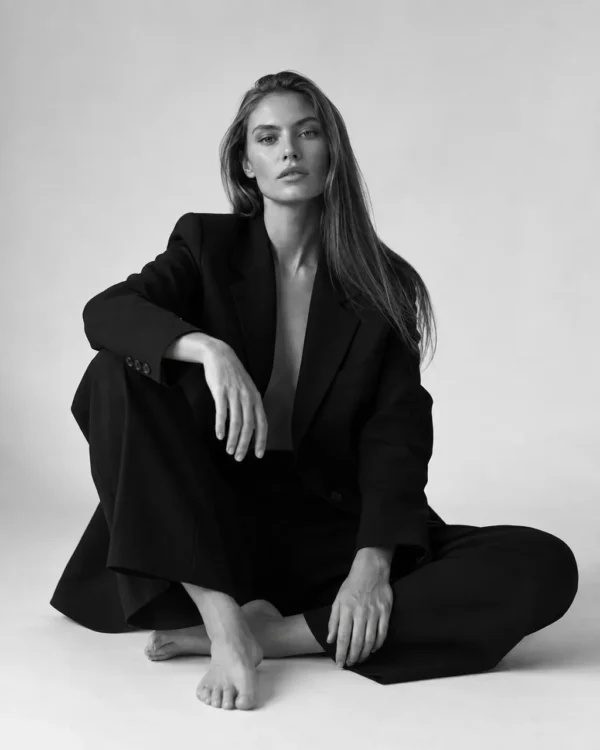 Model in an oversized black blazer and trousers seated barefoot on a studio floor, editorial fashion portrait