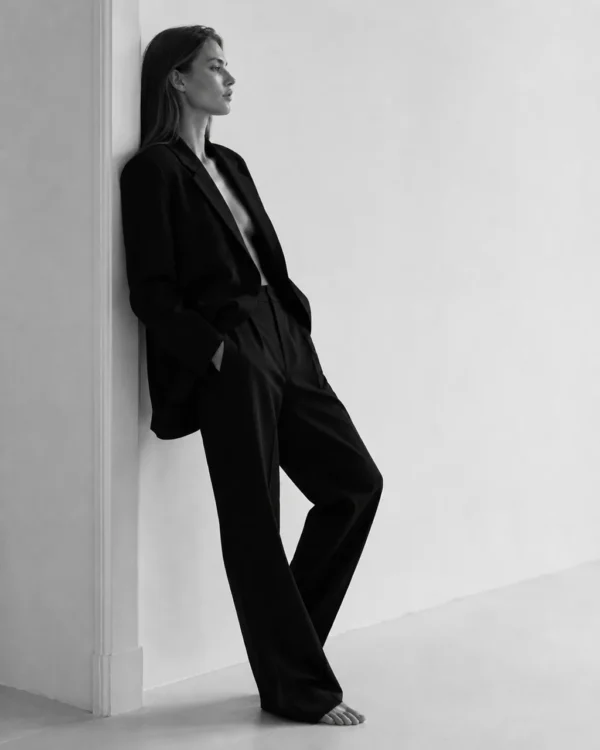 Model in a relaxed black trouser suit leaning against a white wall, full-length editorial fashion shot