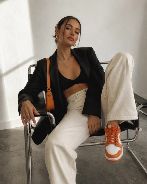 Influencer seated in cream trousers and a black blazer with orange sneakers against concrete