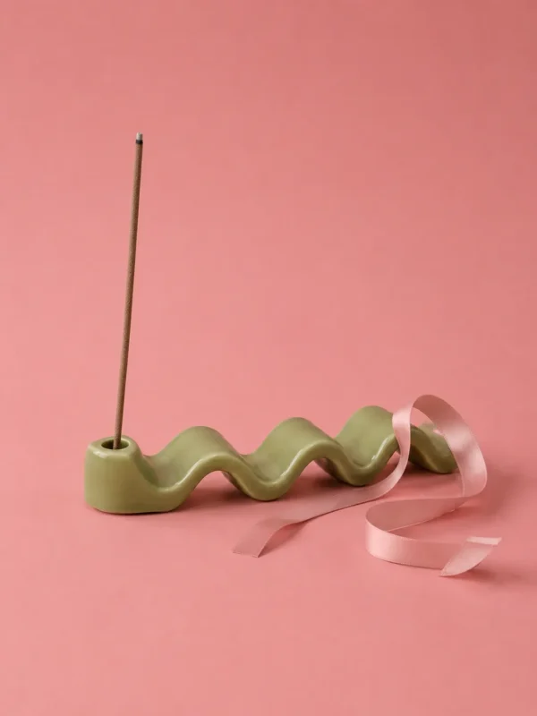 Green ceramic wave-shaped incense stick holder with a pink ribbon on a pink background