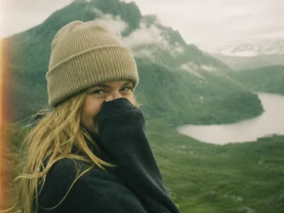Portrait of a hiker in a beanie pulling a collar over her face on a windy fjord ridge on an adventure trip