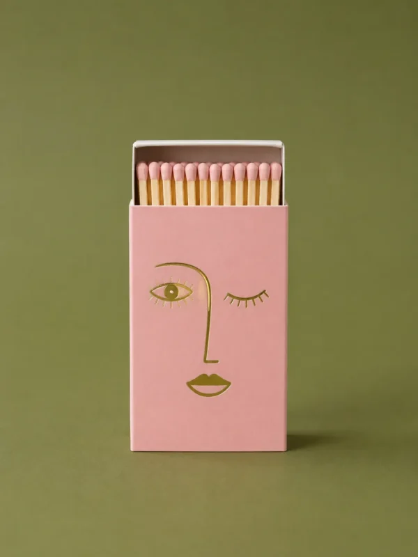 Open pink matchbox printed with a gold line-drawn face, full of white-tipped matches, on olive