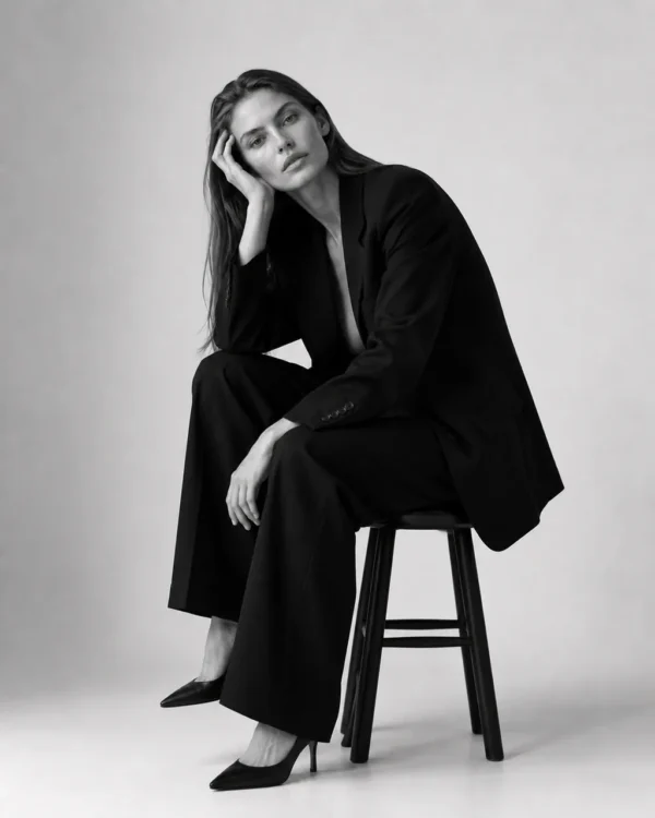 Model in a black suit seated on a stool resting her head on one hand, contemplative editorial fashion portrait