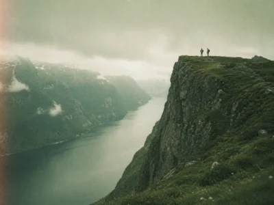 Two small figures standing at the edge of a sheer cliff above a long misty fjord on an adventure trip