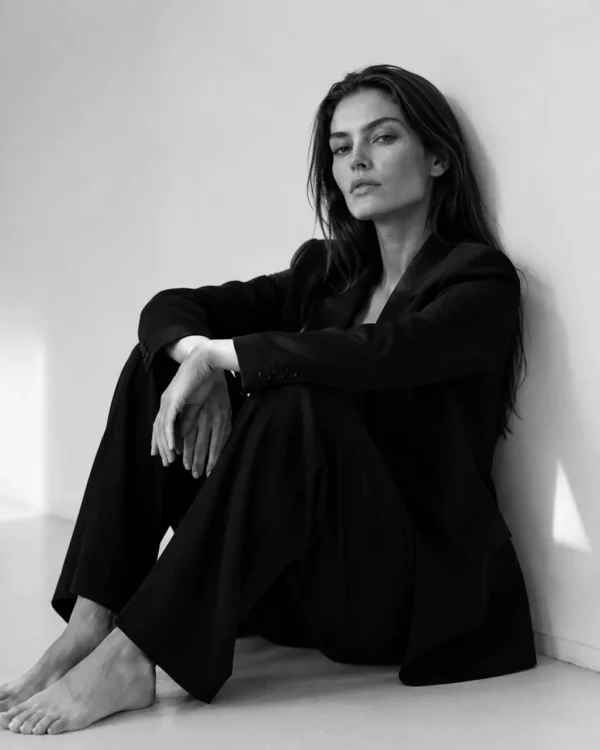 Model in a black suit seated on the floor with one knee up against a bright wall, editorial fashion portrait