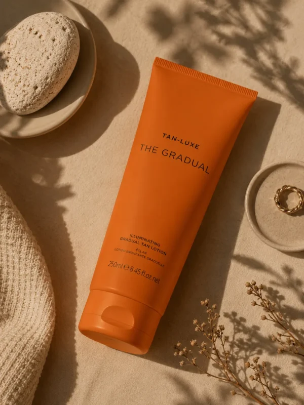 Orange skincare tube on linen with straw hat and leaf shadows in warm light