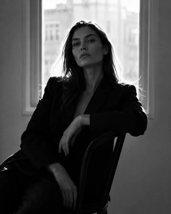 Model in a black suit seated by a large window with a city skyline behind, editorial fashion portrait