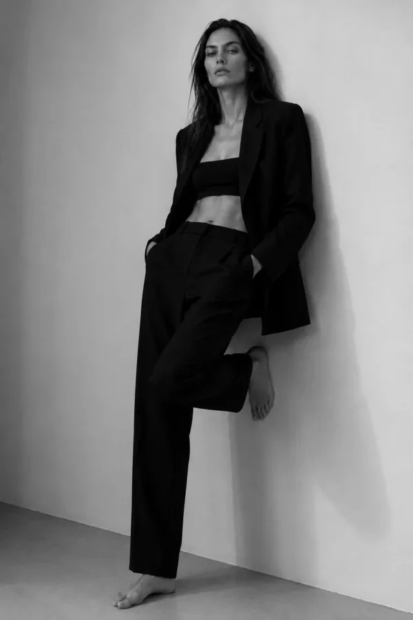 Model in a black suit and bralette standing against a white wall with one foot raised, editorial fashion