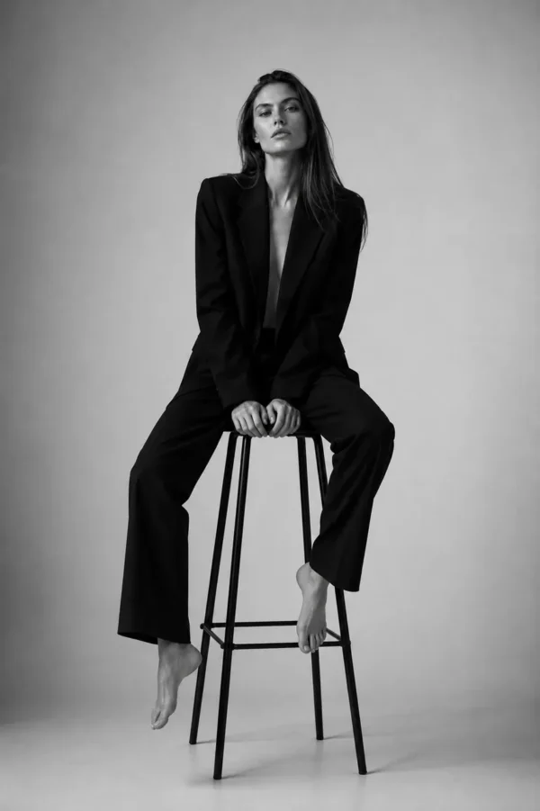 Model in a black suit perched on a tall metal stool in a bright studio, full-length editorial fashion shot