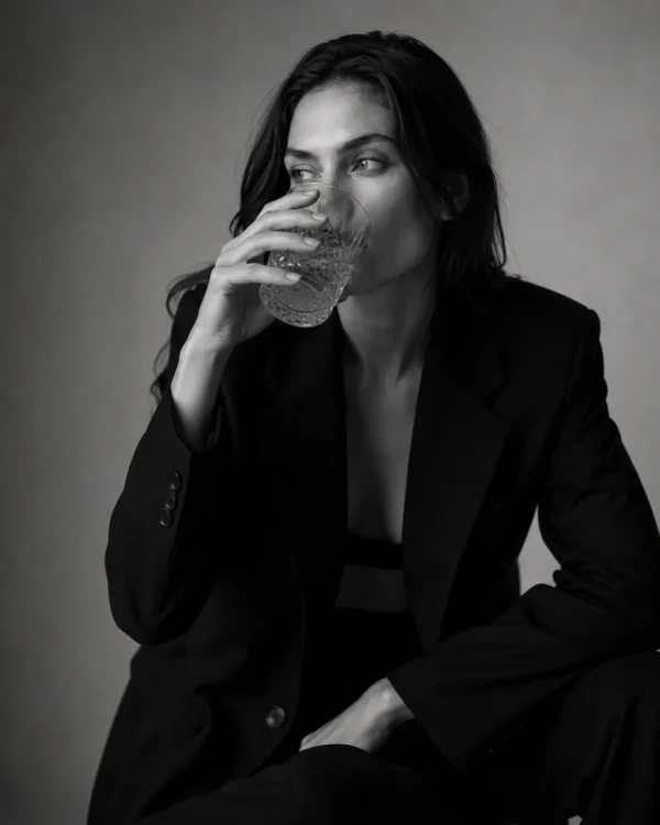 Model in a black suit sipping from a glass against a grey backdrop, candid editorial fashion portrait