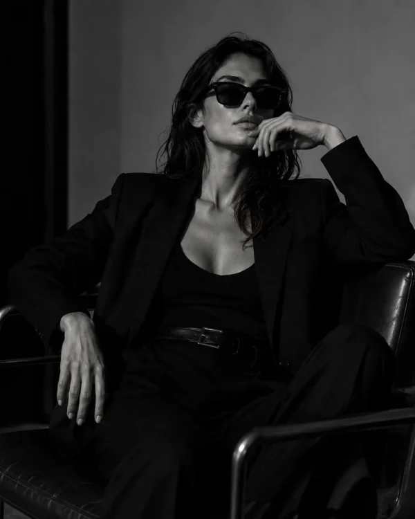 Model in a black suit and sunglasses seated in a leather chair, cool editorial fashion portrait