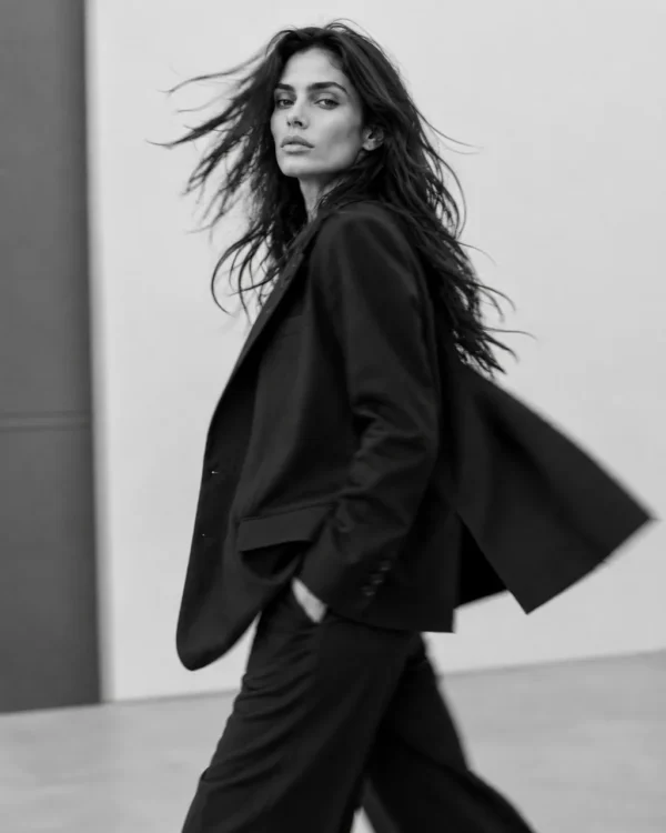 Model in a black blazer caught mid-stride with hair in motion across a bright studio, editorial fashion