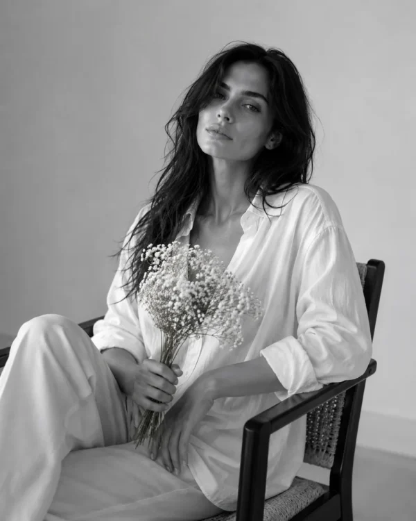 Model in a soft white suit seated on a wooden chair holding dried flowers, editorial fashion portrait