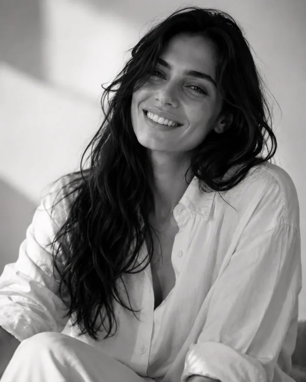 Model in an oversized white shirt smiling with tousled hair in bright light, natural editorial fashion portrait
