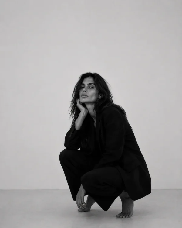 Model in a black suit crouching low against a pale studio wall, editorial fashion full-length portrait